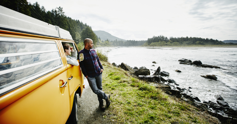 a man in a blue jumper leans against a yellow van as he looks out across a river. Another person sits in the vehicle and looks out the window