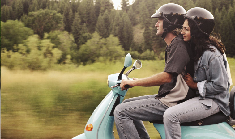 a man and woman sit on a blue scooter as they ride through green country side